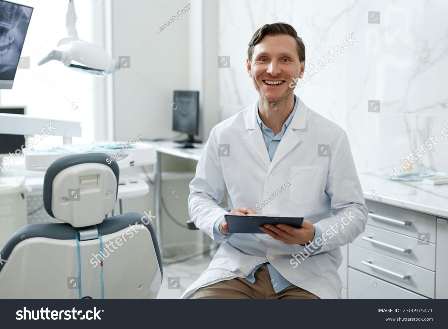 Portrait of smiling male dentist looking at camera in dental clinic interior and holding clipboard, copy space #2300975471