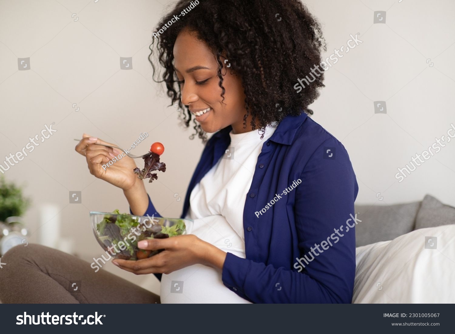 Smiling millennial african american lady with big belly eating salad in room interior, close up. Health care, proper nutrition, vegan and expectation of child, motherhood and family #2301005067