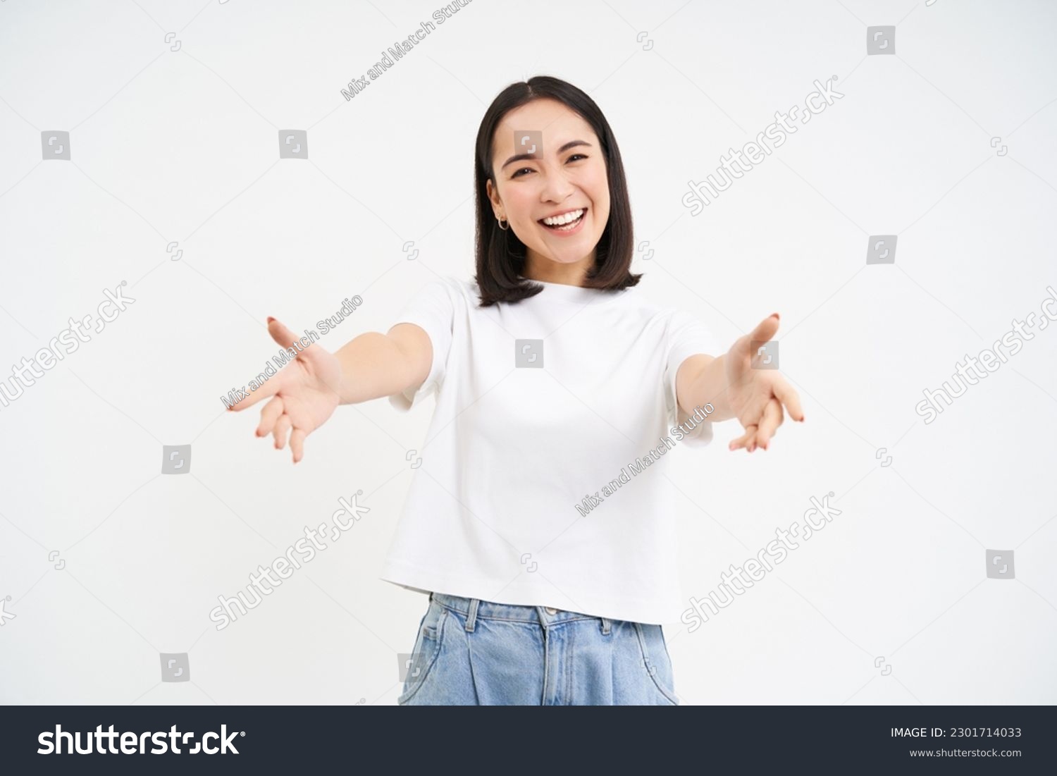 Friendly smiling asian woman, reaching her hands towards camera, hugging, welcoming you, standing over white background. #2301714033