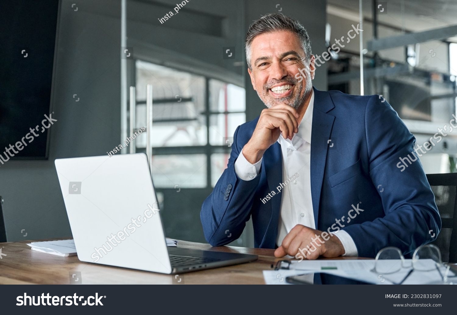 Happy smiling middle aged professional business man company executive ceo manager wearing blue suit sitting at desk in office working on laptop computer laughing at workplace. Portrait. #2302831097