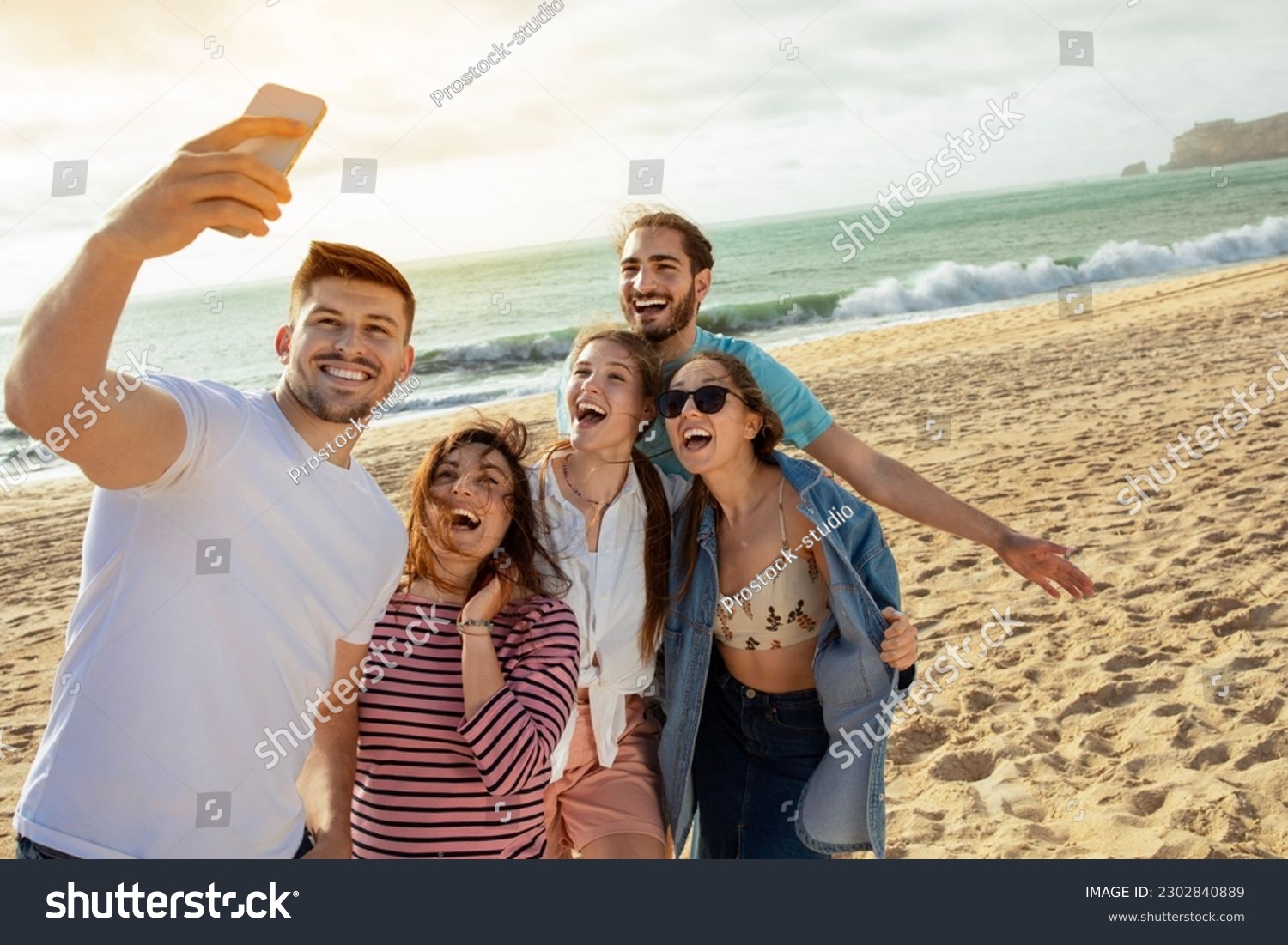 Happy millennial european and arabic friends have fun, taking selfie on smartphone, enjoy free time on ocean beach. App for blog and social network, outdoor party, holiday and trip #2302840889
