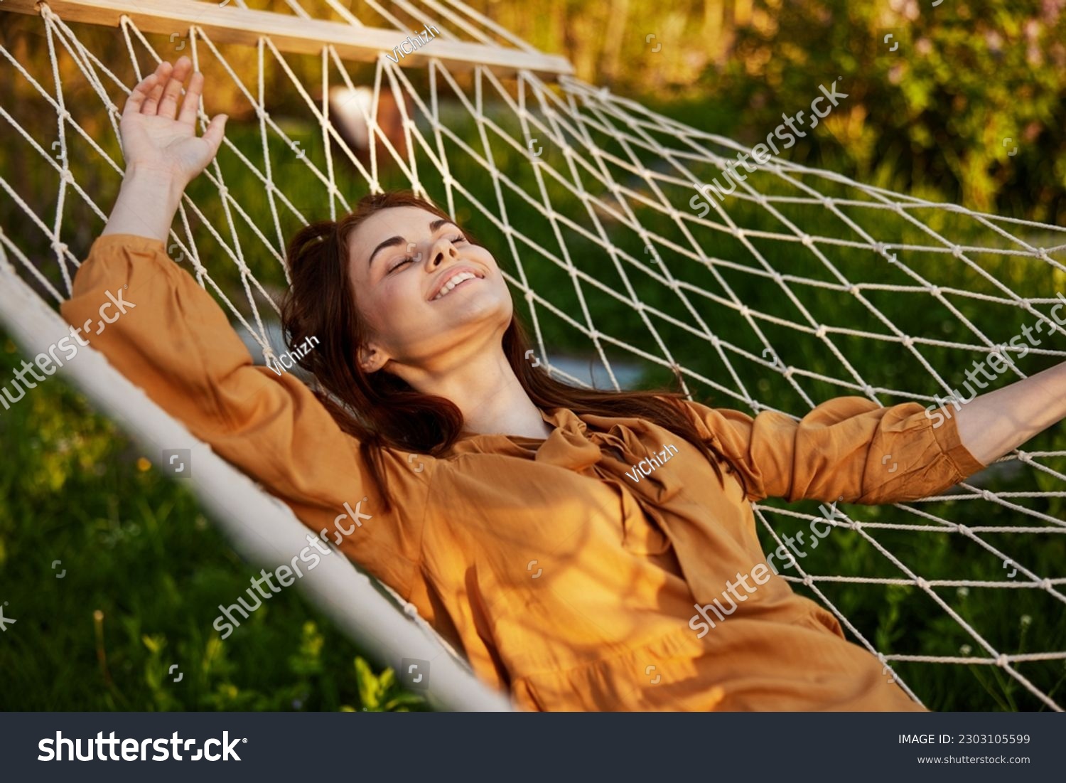a happy woman is resting in a hammock with her eyes closed and her hands behind her head smiling happily enjoying the day #2303105599