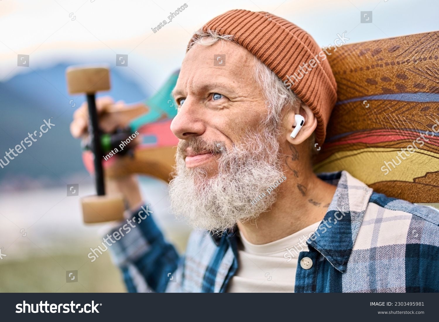 Active cool bearded old hipster man standing in nature park holding skateboard wearing earbud. Mature traveler skater enjoying freedom spirit and extreme sports hobby listening music in earphones. #2303495981