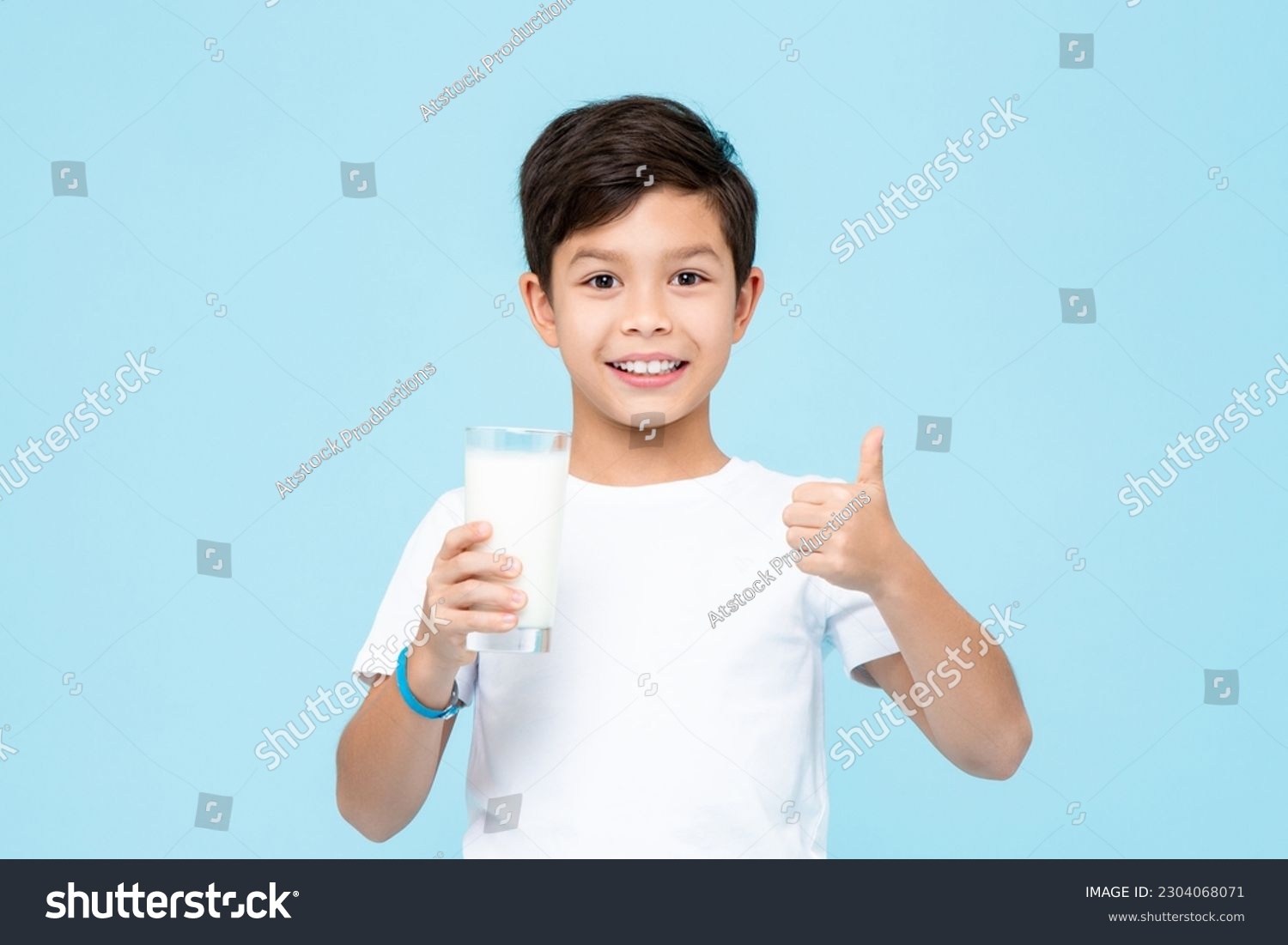 Happy smiling mixed race Asian kid boy holding a glass of fresh milk and giving thumbs up in isolated light blue color background studio shot #2304068071
