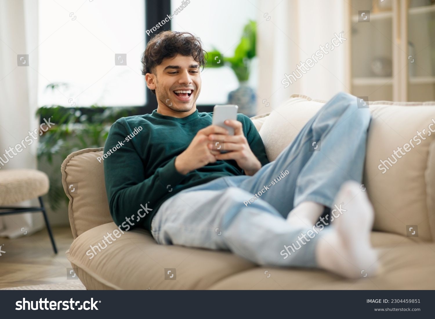 Happy Middle Eastern Young Man Using Cellphone And Laughing Texting Lying On Sofa At Home. Cheerful Man Holding Smartphone Playing Game And Networking Online Having Fun On Weekend #2304459851