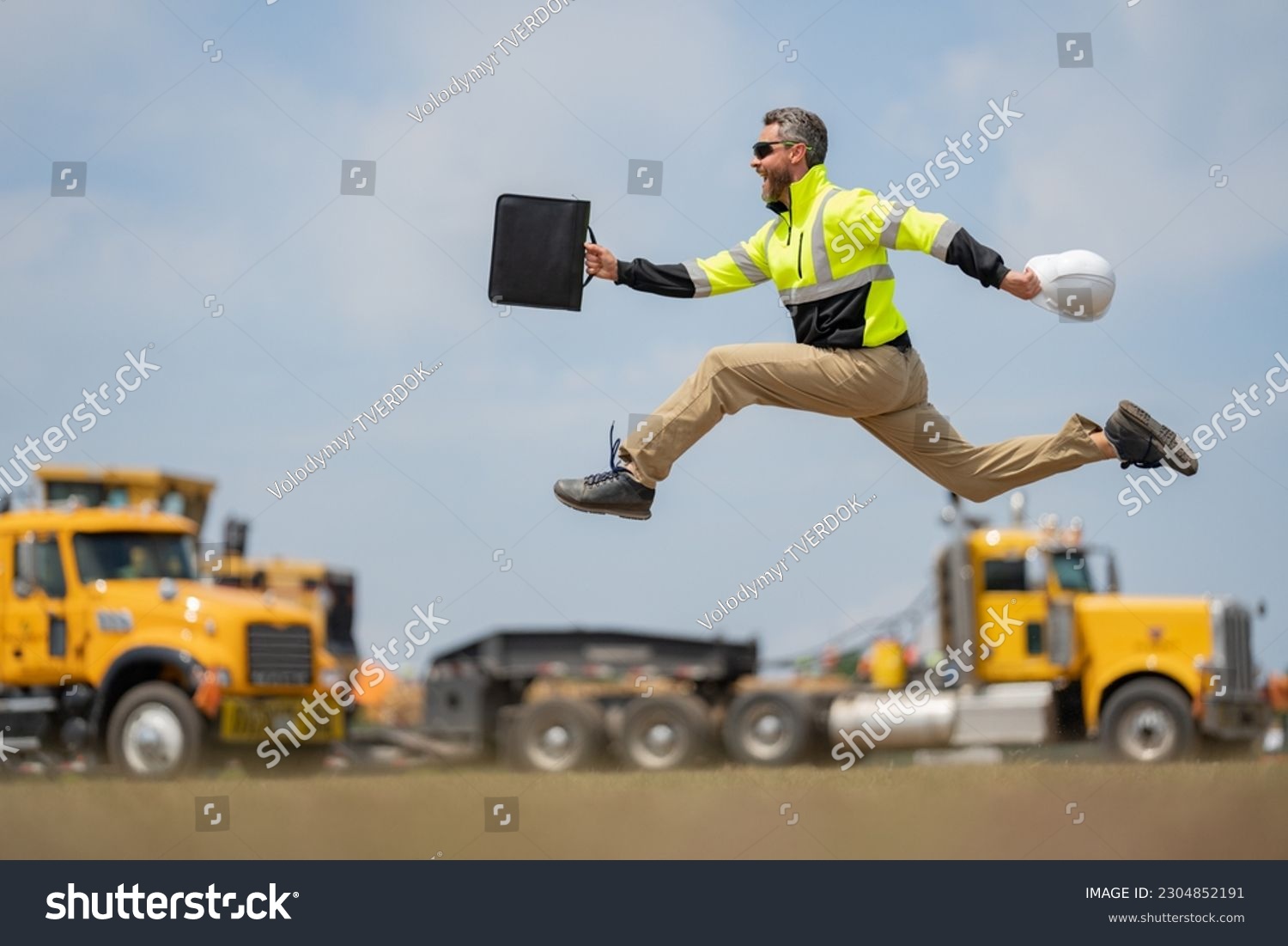 Hispanic 40 s builder excited jump on site construction. Excited builder construction worker in a safety helmet jumping in front of the trucks. Excited crazy builder man in helmet jump outdoor. #2304852191