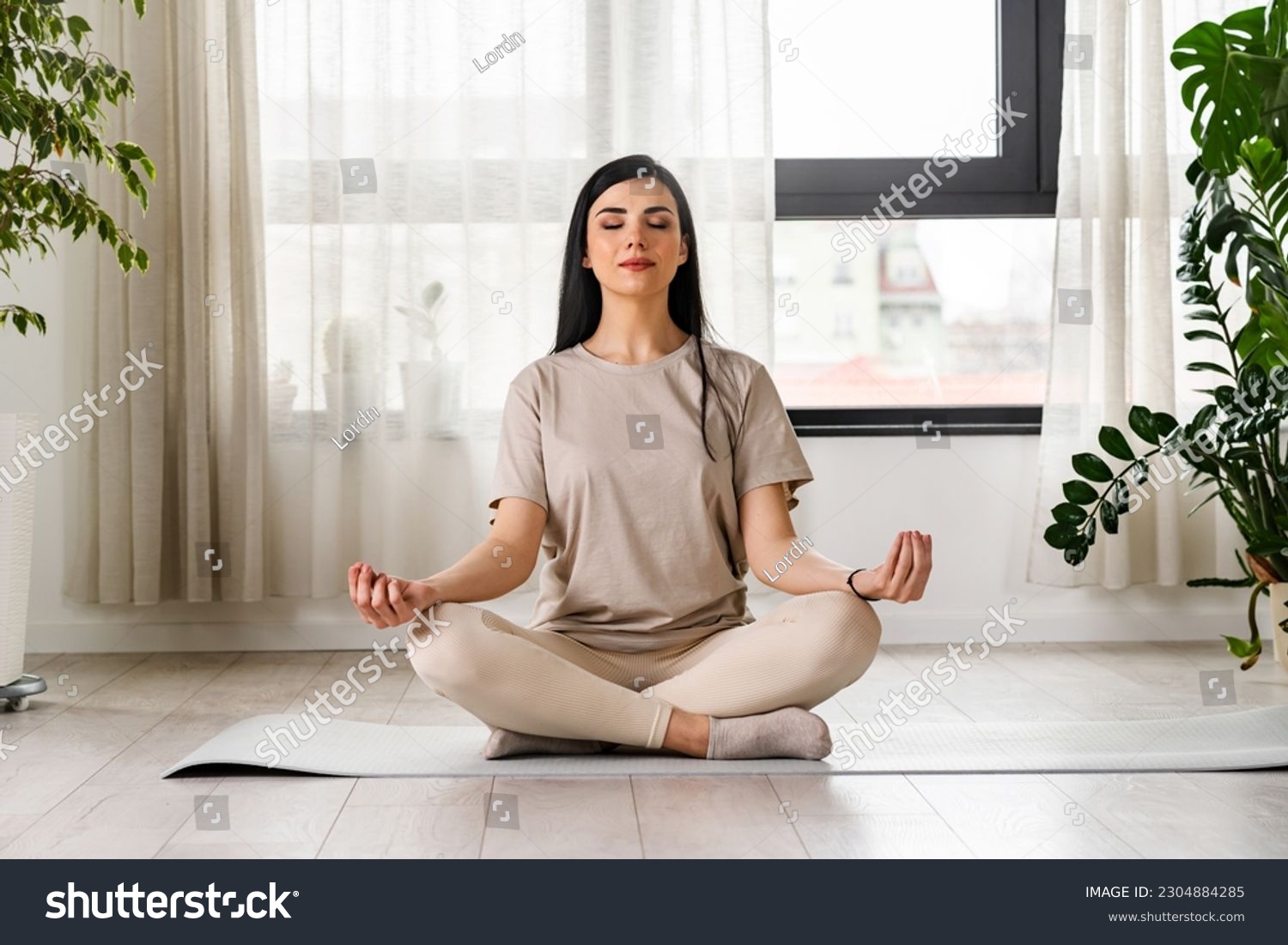 Young woman meditating sitting on a yoga mat near a big window #2304884285