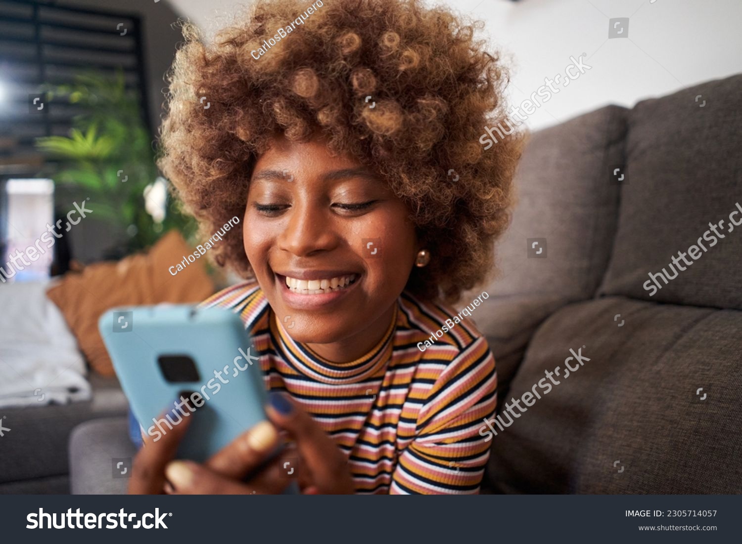 Close up of smiling African American pretty woman using cell phone lying on sofa in living room. Happy young black afro girl flirting on mobile dating app at home. People looking for life partner. #2305714057