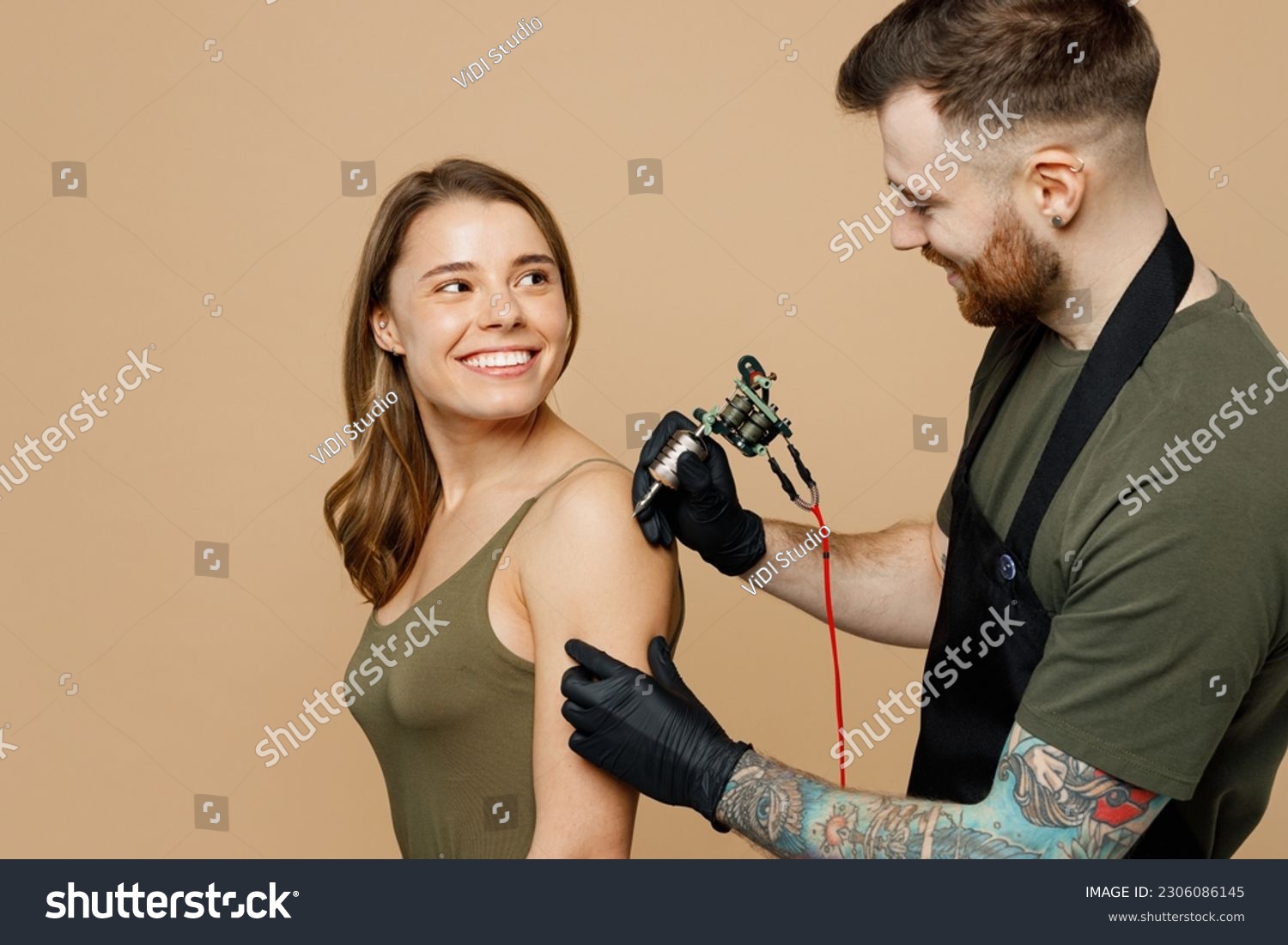 Side view happy tattooer master artist tattooed man wears green t-shirt apron hold machine black ink in jar equipment for making tattoo art on woman female body hand isolated on plain beige background #2306086145