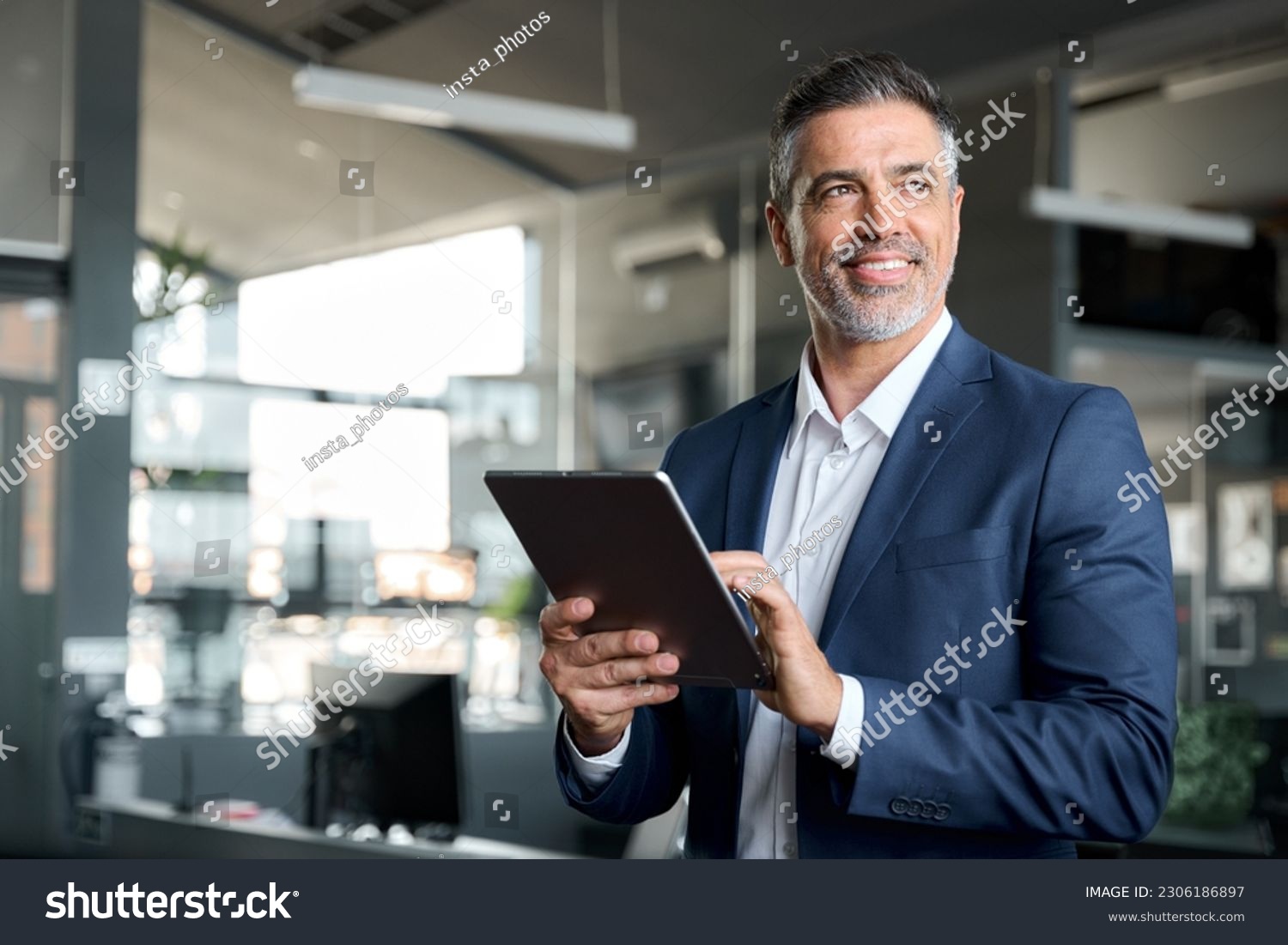 Happy middle aged business man ceo wearing suit standing in office using digital tablet. Smiling mature businessman professional executive manager looking away thinking working on tech device. #2306186897
