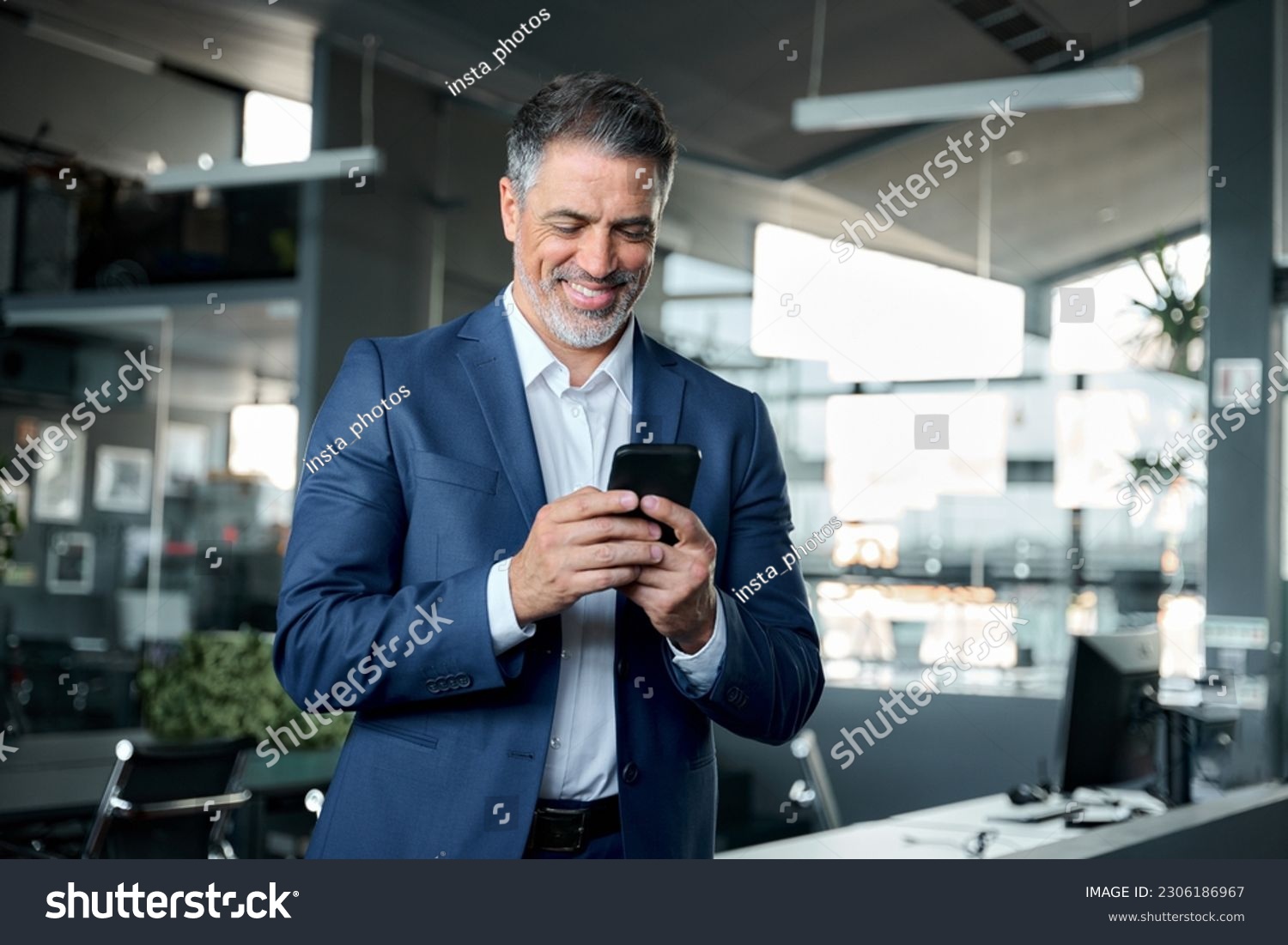 Happy middle-aged business man ceo wearing blue suit standing in office using cell phone. Older businessman professional executive holding mobile satisfied with enterprise solution management service. #2306186967