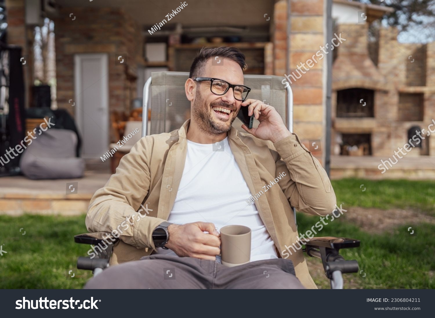 One adult man 40 years old caucasian male sit in front of his house on vacation talk on his smartphone mobile phone call happy smile during the conversation real person copy space #2306804211