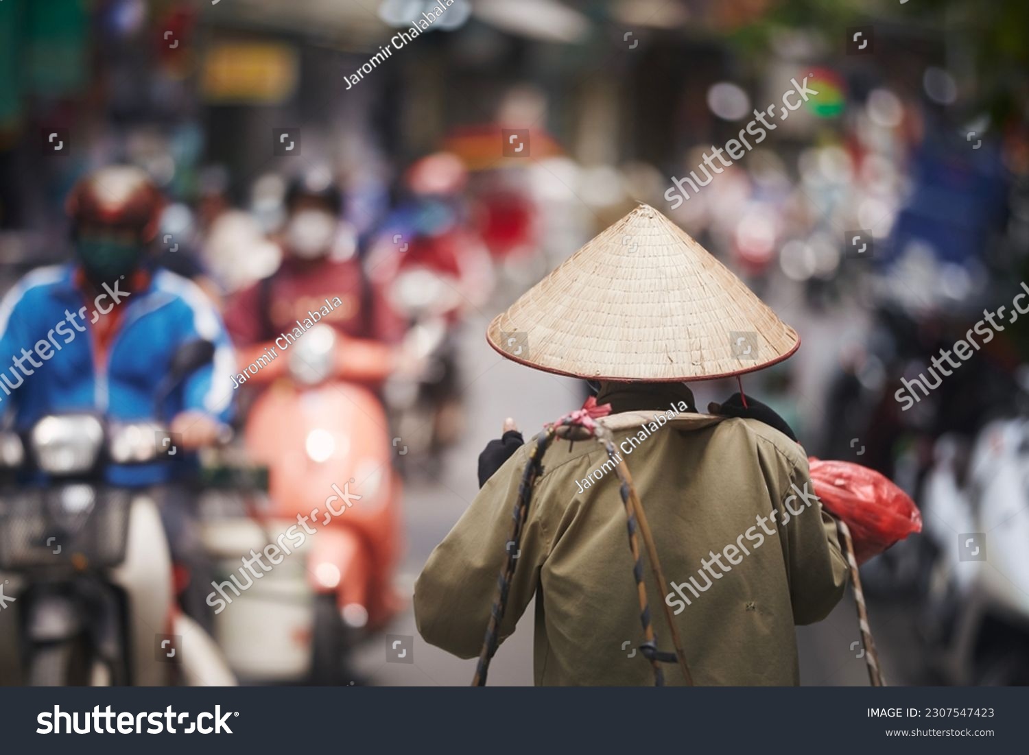 Selective focus on traditional conical hat of person walking against traffic motorbikes on busy street in Old Quarter in Hanoi, Vietnam.
 #2307547423