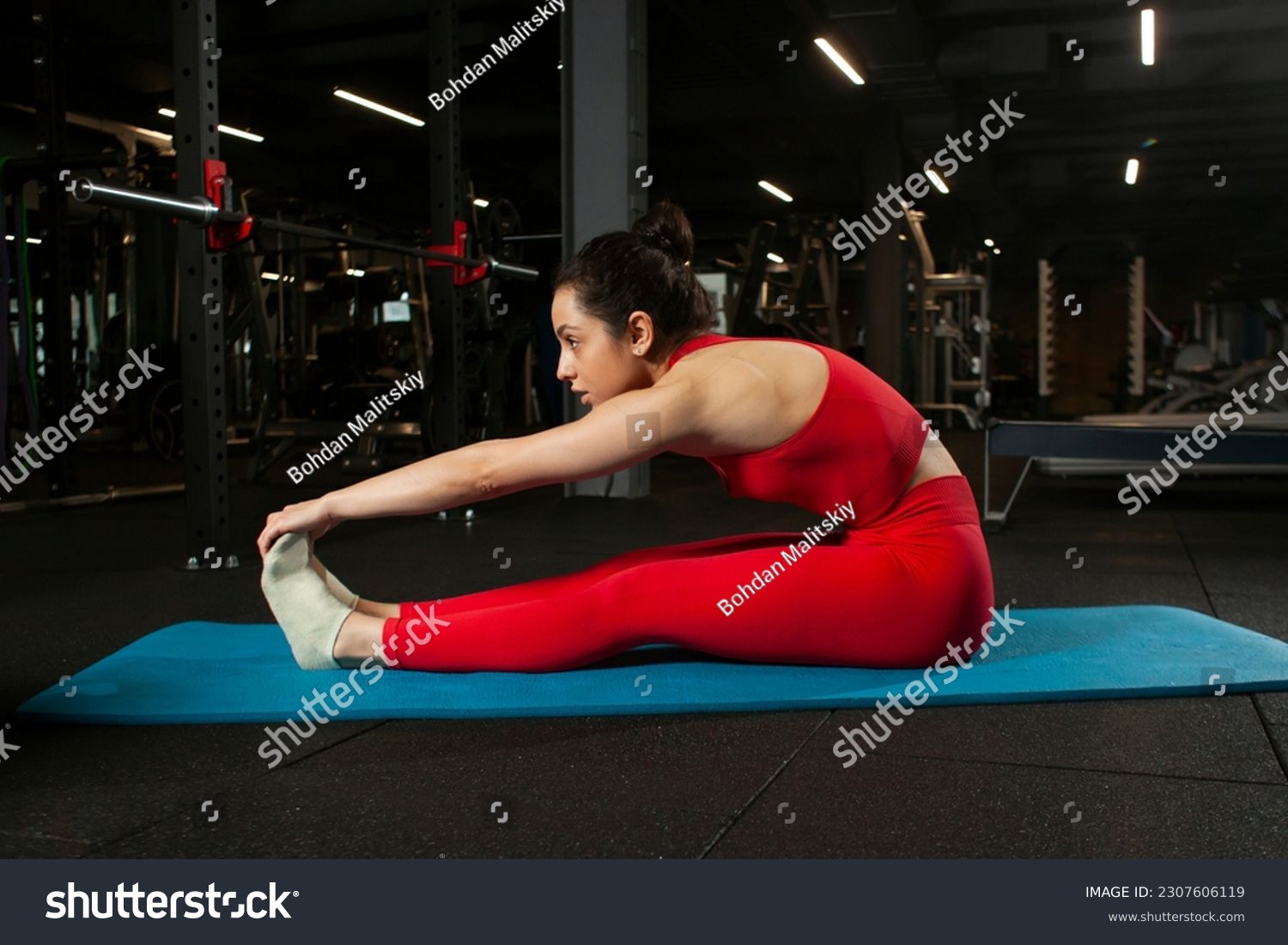 athletic woman in red sportswear sitting on yoga matte in black gym and warming up, girl doing yoga and stretching, attractive woman in fitness club doing flexibility exercise #2307606119