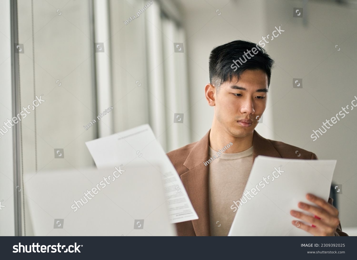 Young busy Asian business man lawyer, tax accountant manager holding paper documents checking bills, doing sales invoice accounting data, reading legal contract or bank statement in office. #2309392025