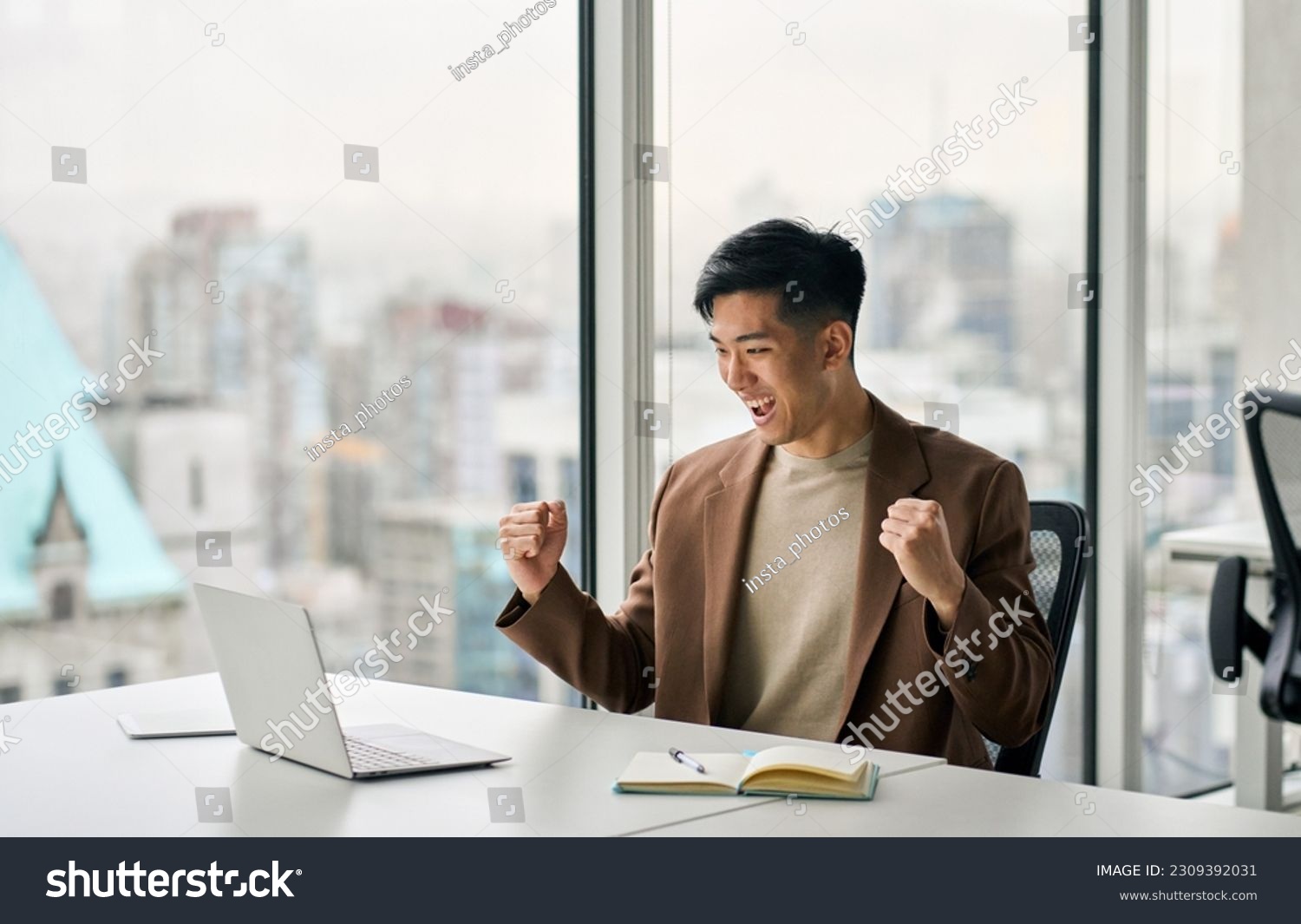 Happy young Asian business man celebrating success at work in office looking at laptop excited about financial trading growth, goals achievement good online results, screaming yes watching game. #2309392031