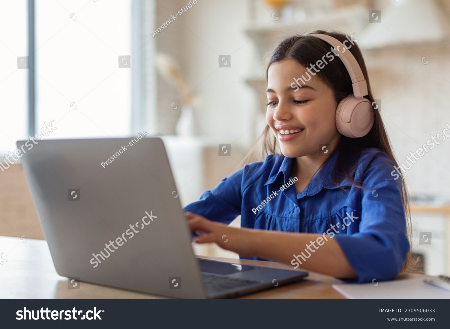 E-Learning Class. Cheerful Arabic Schoolgirl Studying Online Typing On Laptop Computer Wearing Earphones Sitting At Home. School Kid Girl Websurfing Enjoying Distance Lesson #2309506033