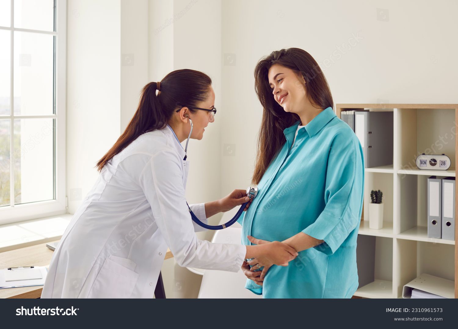 Happy pregnant woman has appointment with doctor at clinic. Female gynaecologist OB GYN medic specialist with stethoscope listens to baby's heartbeat in mother's belly. Pregnancy, health care concept #2310961573