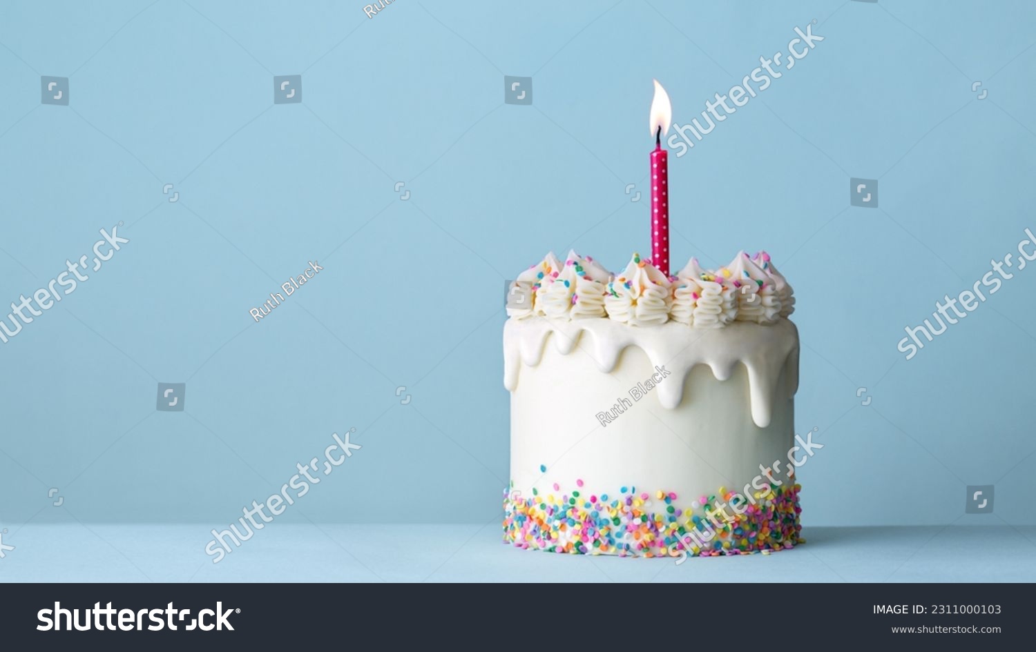 Celebration birthday cake decorated with white drip icing, buttercream frosting swirls, colorful sugar sprinkles and one birthday candle against a plain blue background #2311000103