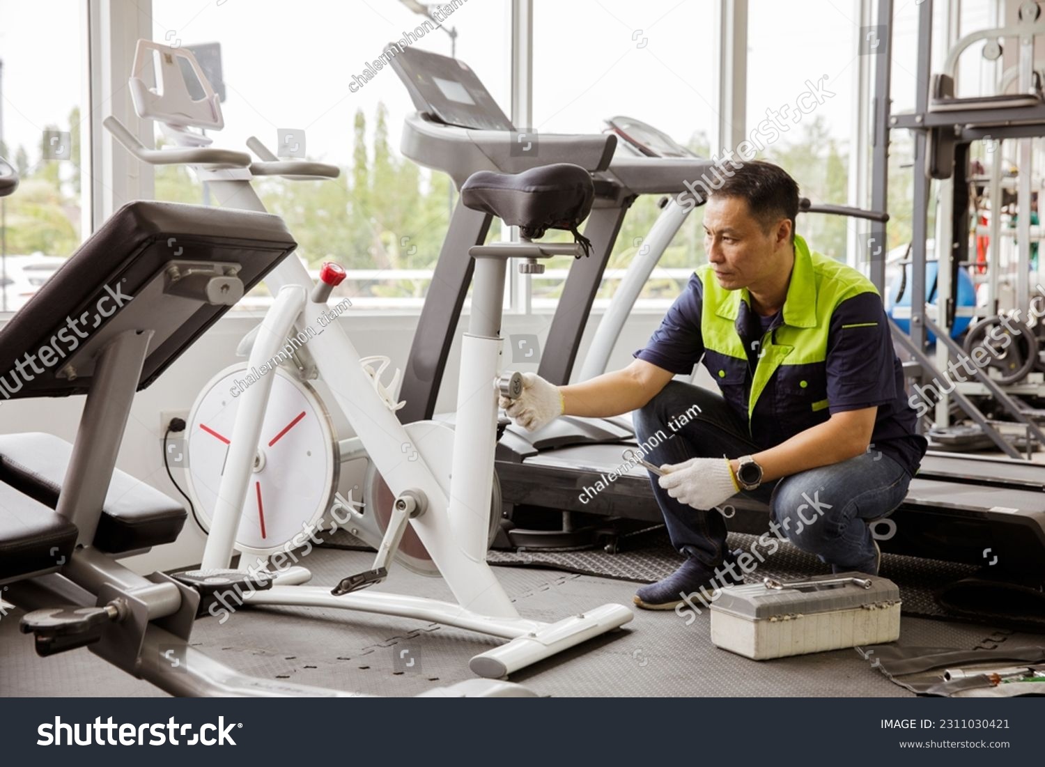 Professional asian male service repair worker or sport fitter using bicycle and treadmill service equipment supervising and securing fitness equipment in indoor gym provide security safety for users. #2311030421