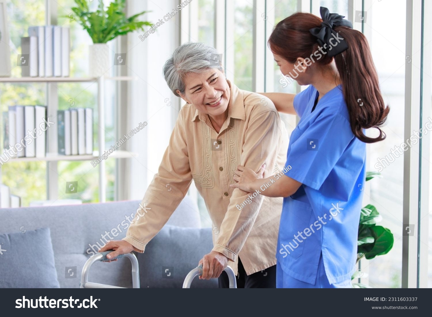Asian young professional successful friendly female nurse in blue hospital uniform helping supporting physical therapy senior old pensioner unhealthy injury woman patient walking via four legs walker. #2311603337