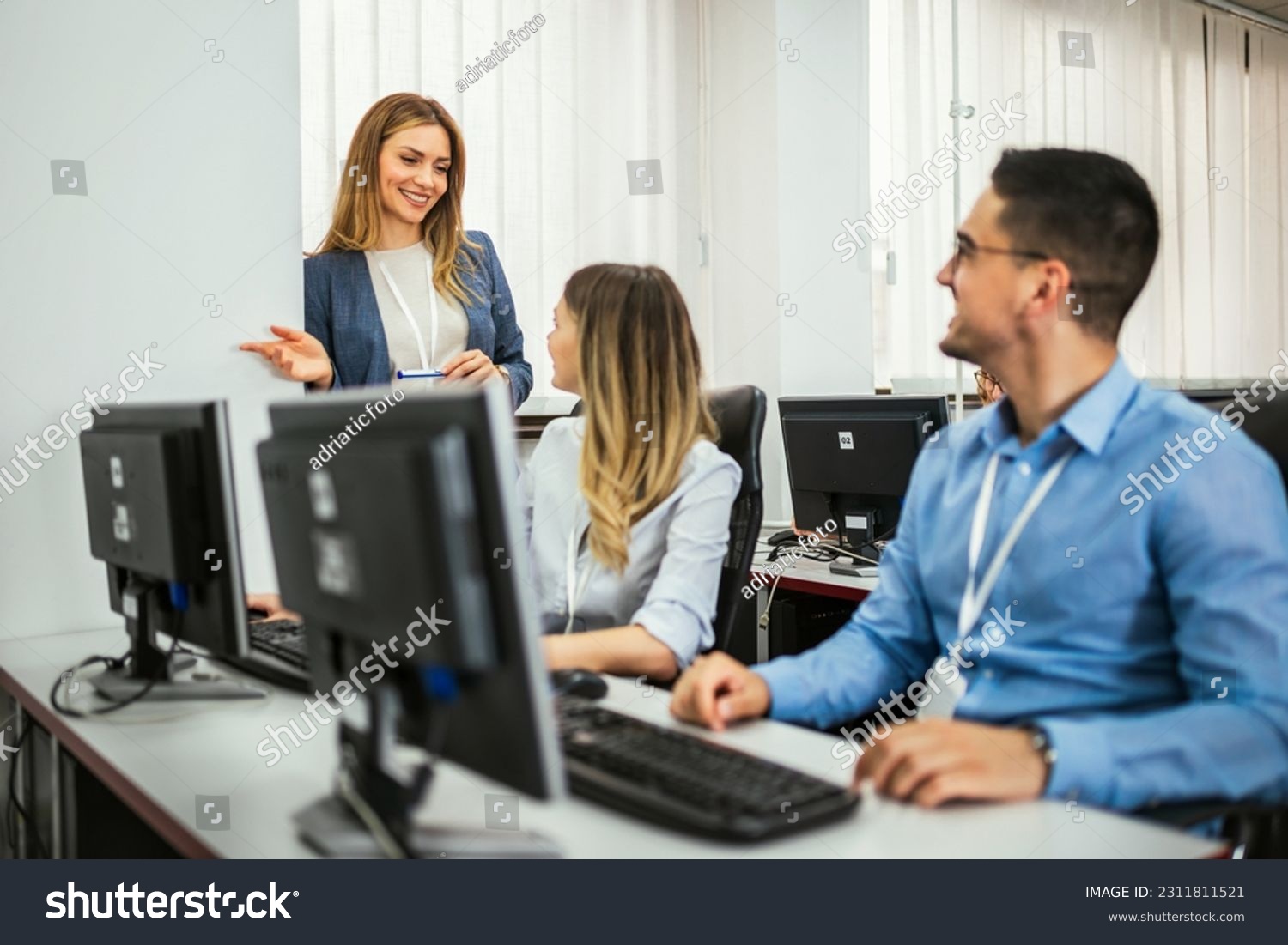 A group of students each sit in front of a desktop in a computer lab. They are each focused on their screens as their female teacher makes her way around to check on each student individually. #2311811521