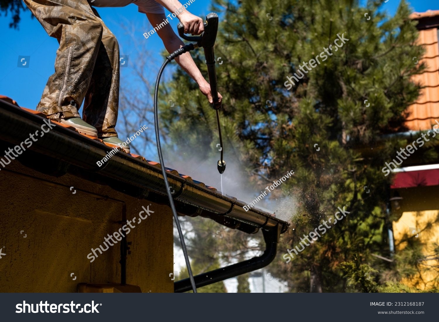 Worker standing on the roof and cleaning rain gutter with high pressure water jet. Professional with equipment for roof gutter cleaning. Maintenance and housekeeping concept of drainage channel. #2312168187