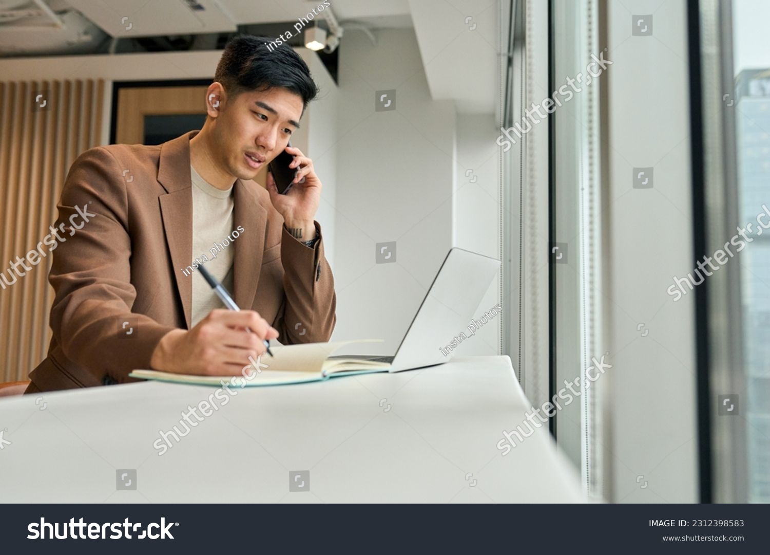 Busy Asian young business man sales manager making phone call using laptop in office. Japanese businessman talking on cellphone working consulting client discussing online digital financial data. #2312398583
