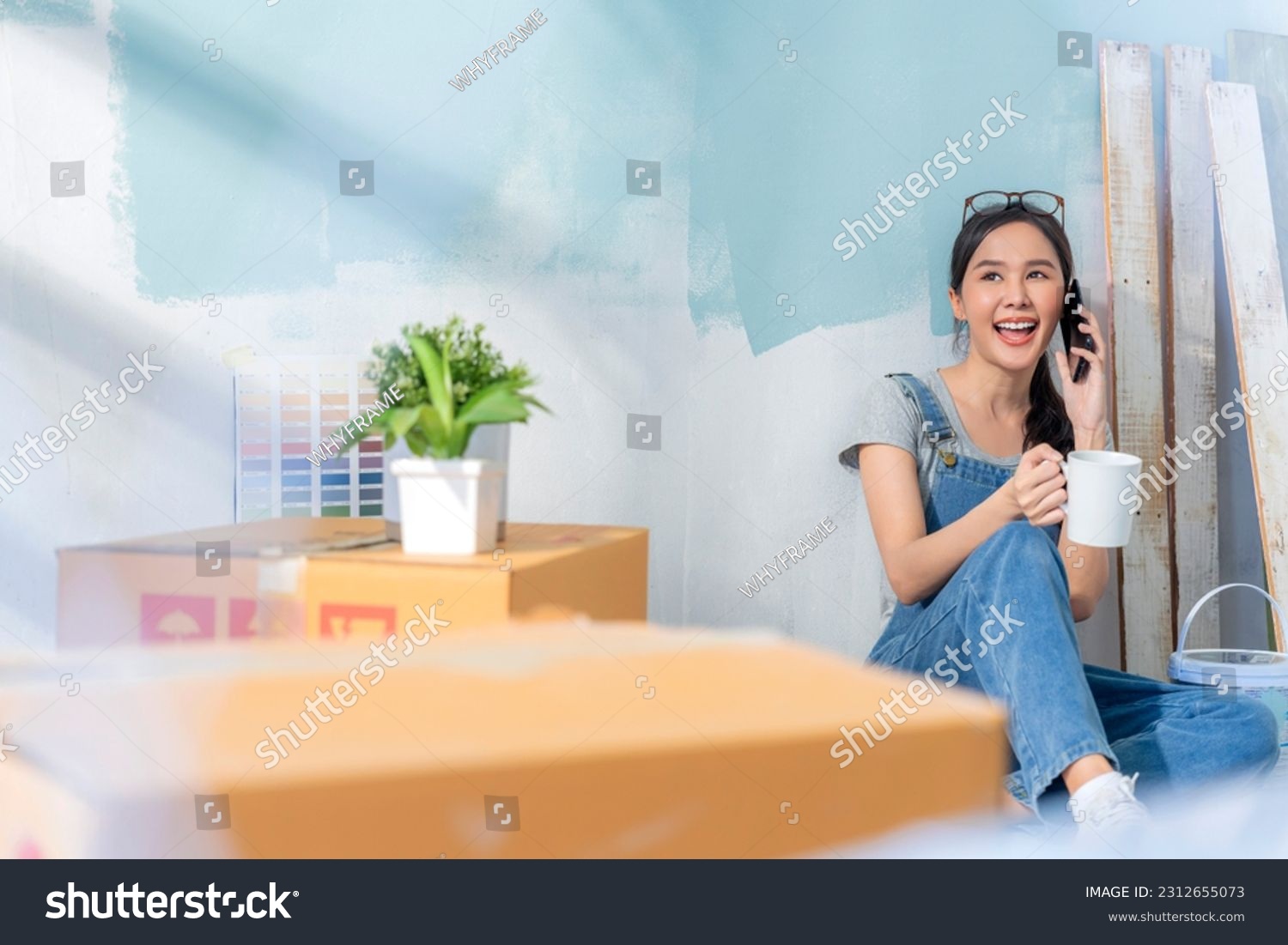 Happy Asian attractive young woman having coffee break during home renovation her living room. Smiling asian woman smartphone conversation to her friend while waiting her new wall colour is ready #2312655073