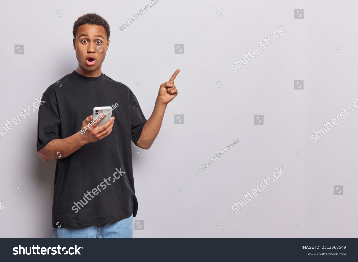 Studio shot of shocked speechless man stares with omg expression at camera points index finger at blank space holds mobile phone dressed in casual black t shirt and jeans isolated on white background #2312668549