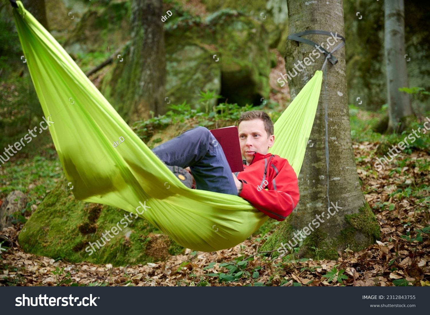 An adventurer rests in a hammock. After the expedition, the tourist rests in a hammock, reads a book in the forest, basking in the warmth of a sunny day. #2312843755