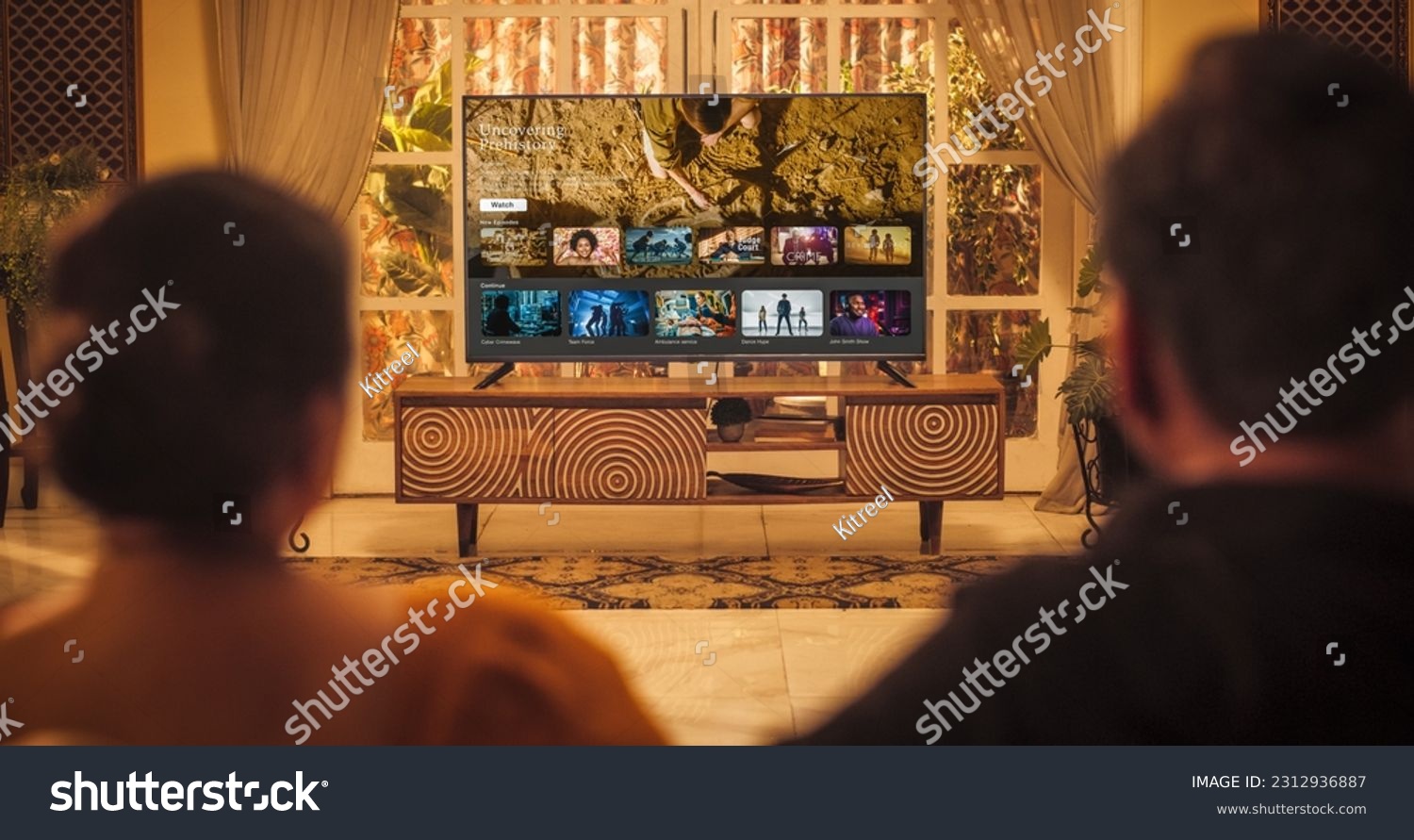 Back View of Elderly Indian Couple Watching TV while Sitting on a Couch in the Living Room. Relaxed Evening for Enjoying Favourite Movies, TV Shows, Content and Ads Thank to Streaming Online Service #2312936887
