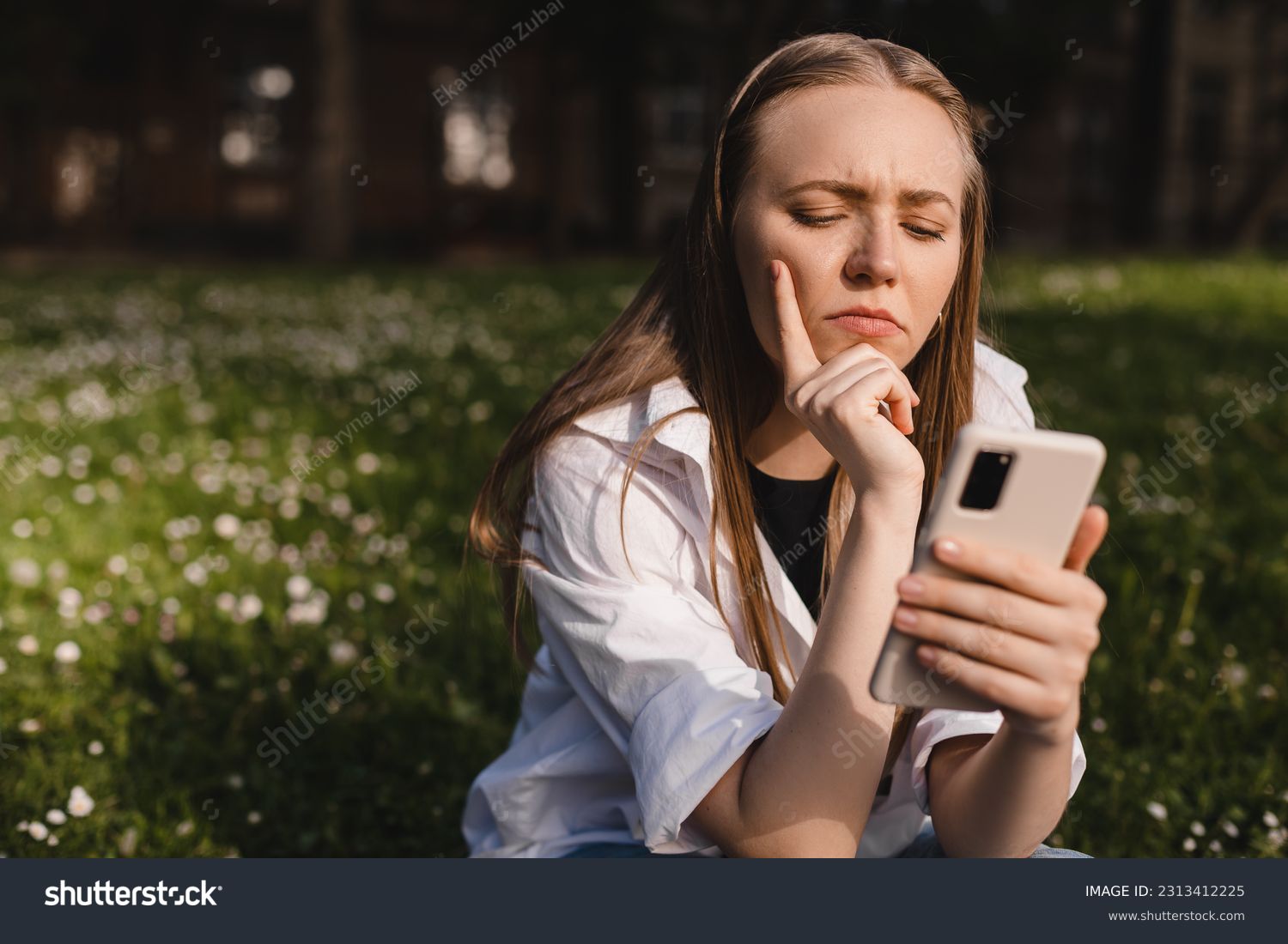 Woman holding a phone while holding her hand on chin. Expressive student thinking outside in campus - Woman in doubt with smartphone in her palm, think about reading information, search solution. #2313412225