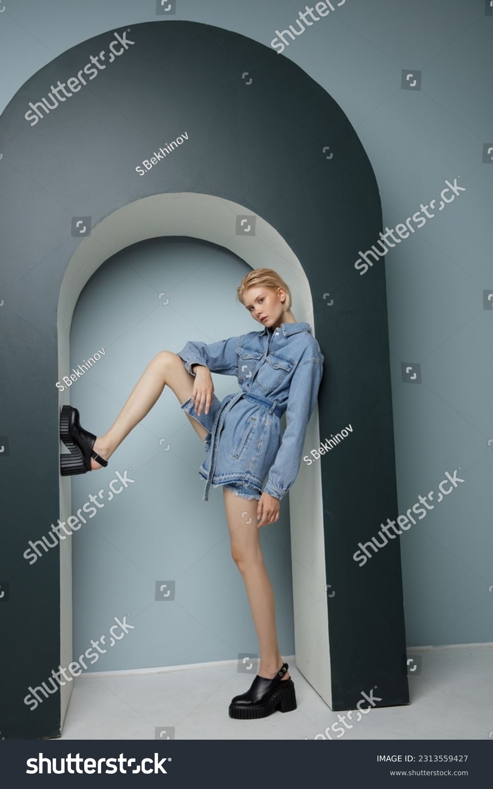 High fashion photo of a beautiful elegant young woman in pretty blue denim, jeans jumpsuit. Bicolor wall, light and dark blue. Blonde, slim figure, short haircut #2313559427