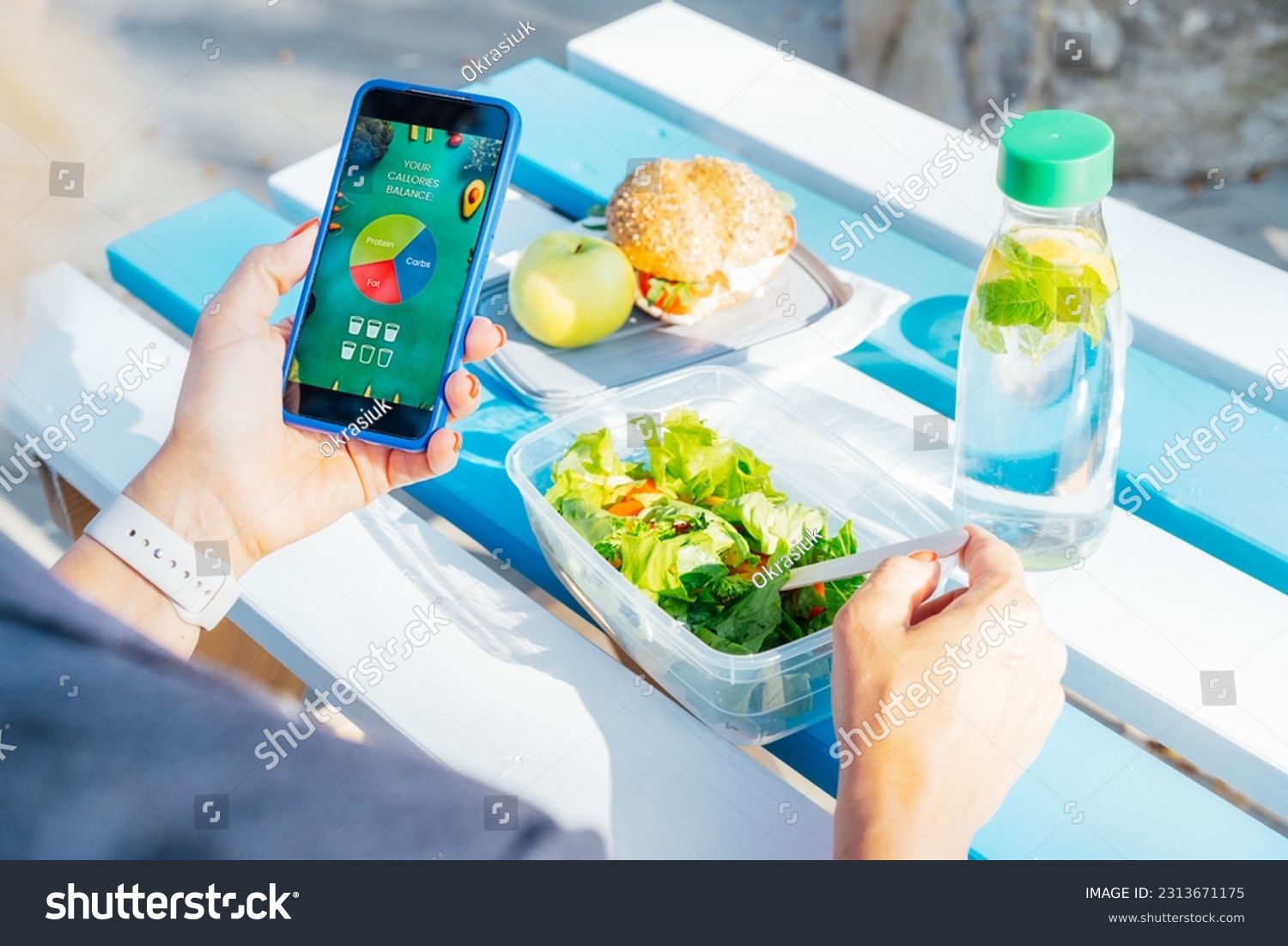 Close up woman using meal tracker app on phone while eating salad at picnic table in the park on a break. Healthy balanced diet lunch box. Healthy diet plan for weight loss. Selective focus #2313671175