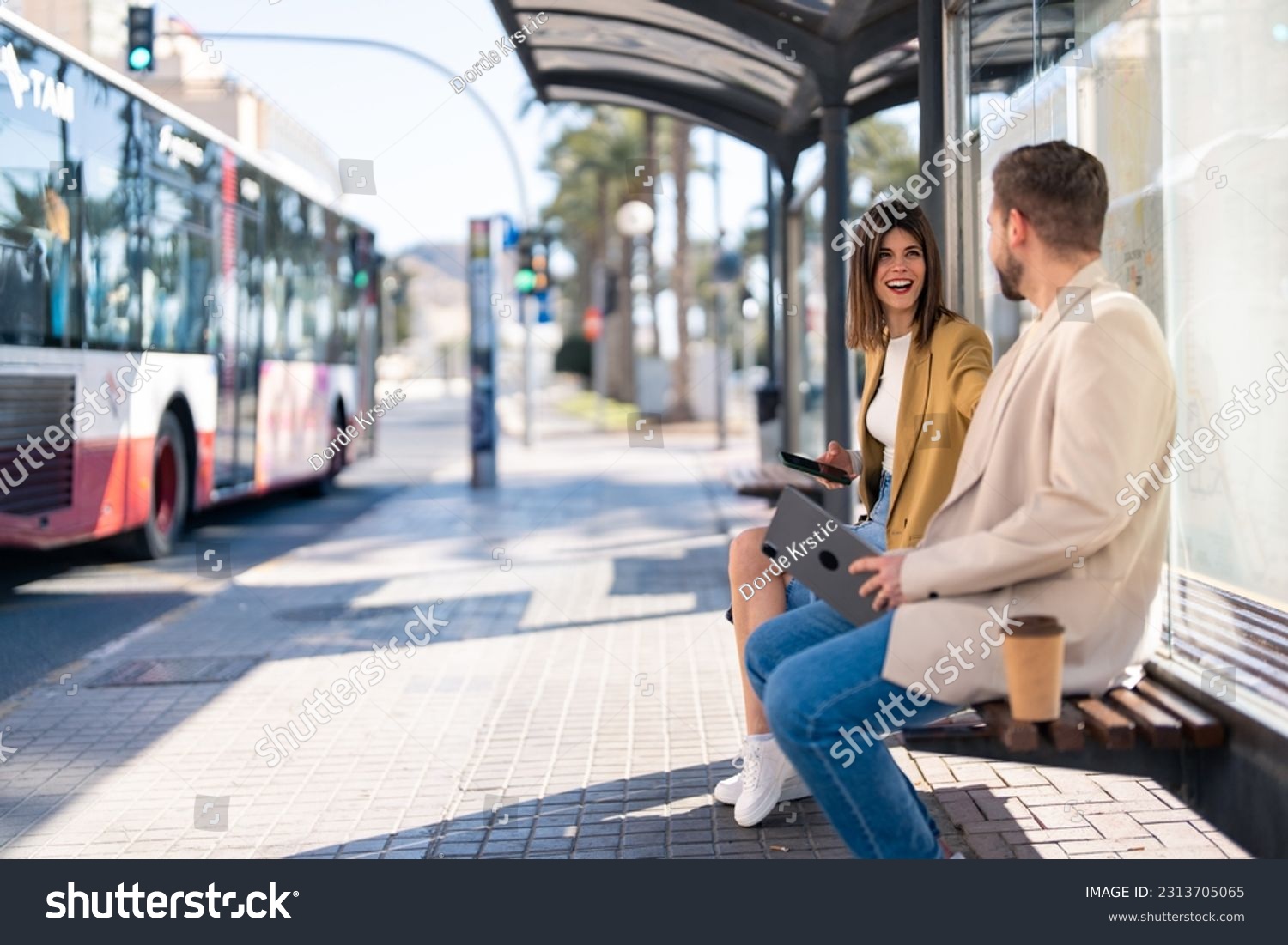 Two cheerful friends young woman and man sitting at bus station while city bus is passing by on the street, having a conversation, laughing, holding mobile phone and digital tablet. Copy space photo. #2313705065