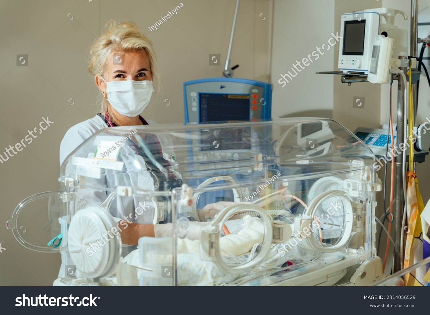 A blonde female doctor in a protective mask caring to a sick baby opening or clothing the incubator. Neonatal intensive care. #2314056529