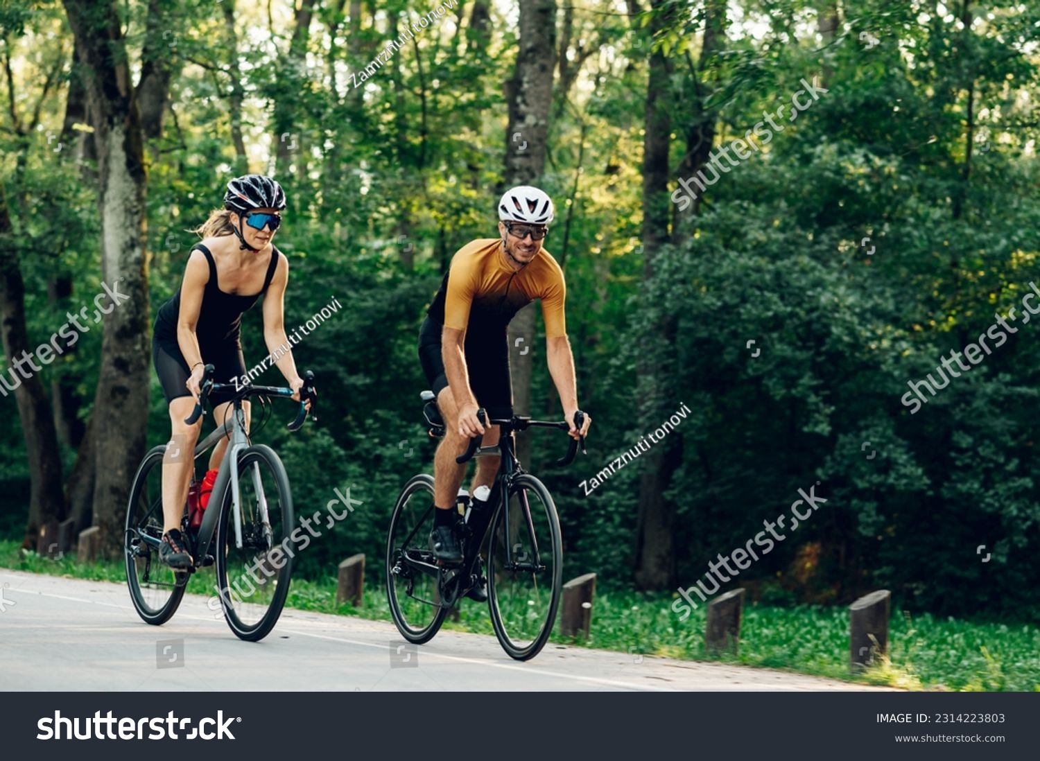 Smiling couple of athletes biking fast on paved road outside of the city. Countryside area, sunny day outdoors. Professional road bicycle racers in action. Concept of endurance and strength. #2314223803
