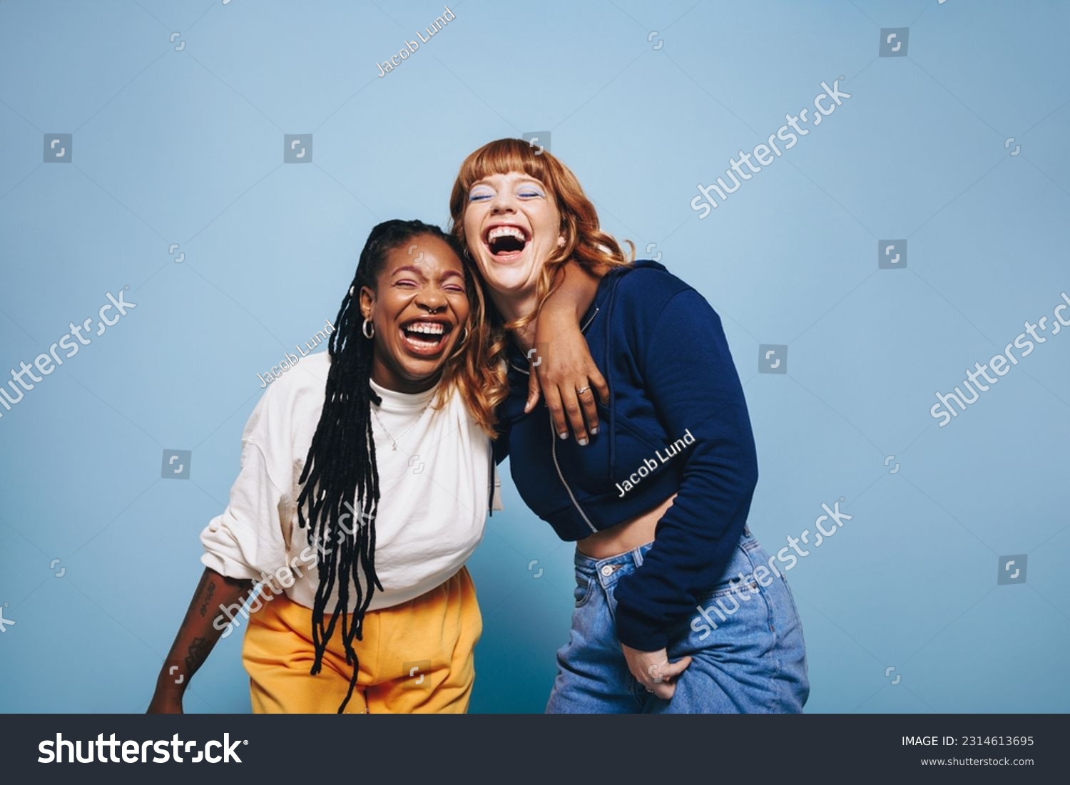 Best friends laughing and having a good time together in a studio. Happy young women enjoying themselves while standing against a blue background. Two vibrant female friends making memories. #2314613695