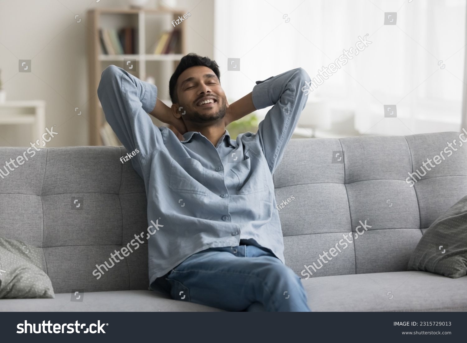 Positive relaxed young Indian man sitting on home sofa, stretching body, leaning on soft back, touching neck with closed eyes, enjoying comfort, coziness, breathing fresh air #2315729013