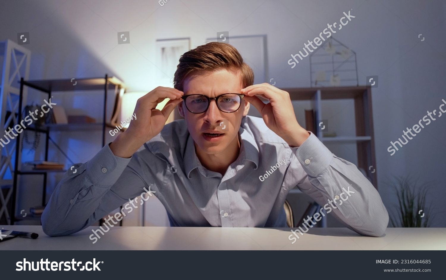 Shocked man. Online conference. Interesting news. Smart curious guy sitting work desk looking on camera holding glasses in light room interior. #2316044685
