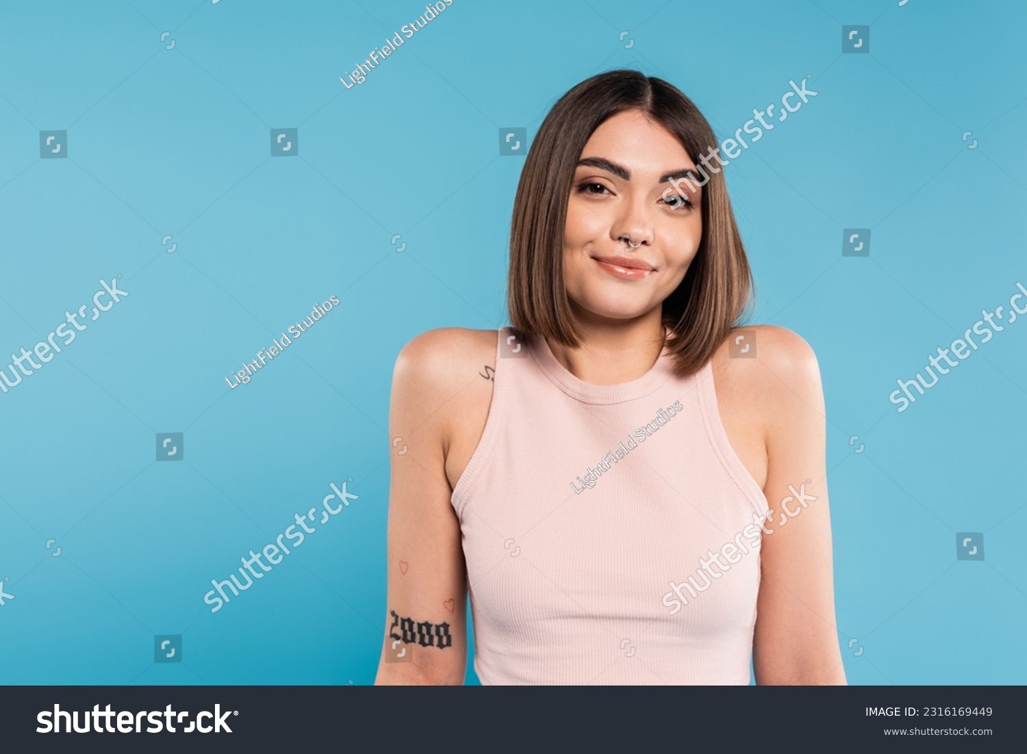 not knowing, smiling young woman with tattoos and nose piercing standing in tank top on blue background, looking at camera, confused, pretty face, generation z, summer outfit #2316169449