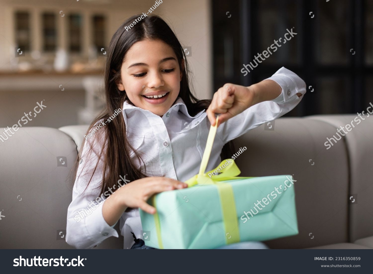 Birthday Gift. Happy Middle Eastern Kid Girl Opening Present Box Celebrating Holiday Sitting On Sofa At Home. Child Posing With Wrapped Gift. B-Day Celebration Concept. Selective Focus #2316350859