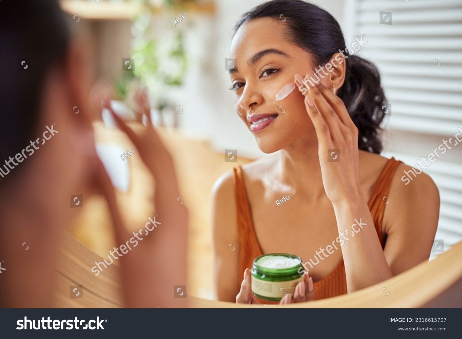 Woman caring of her beautiful skin on the face standing near mirror in the bathroom. Mexican woman applying moisturizer on her face at home. Multiethnic girl holding little green jar of bio skin cream #2316615707