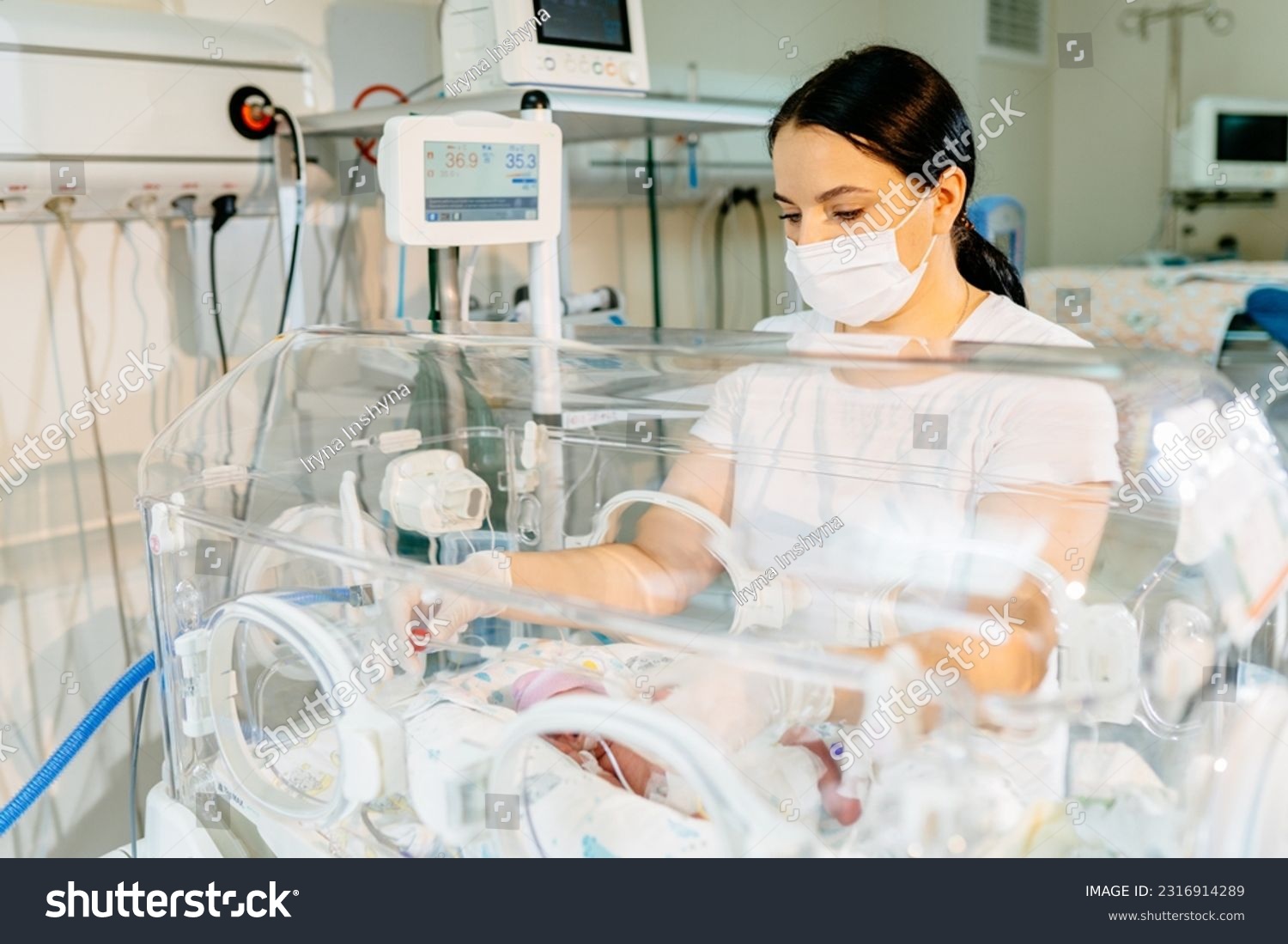 A brunette female nurse in a protective mask caring to a sick premature baby standing next to the incubator. Neonatal intensive care. #2316914289