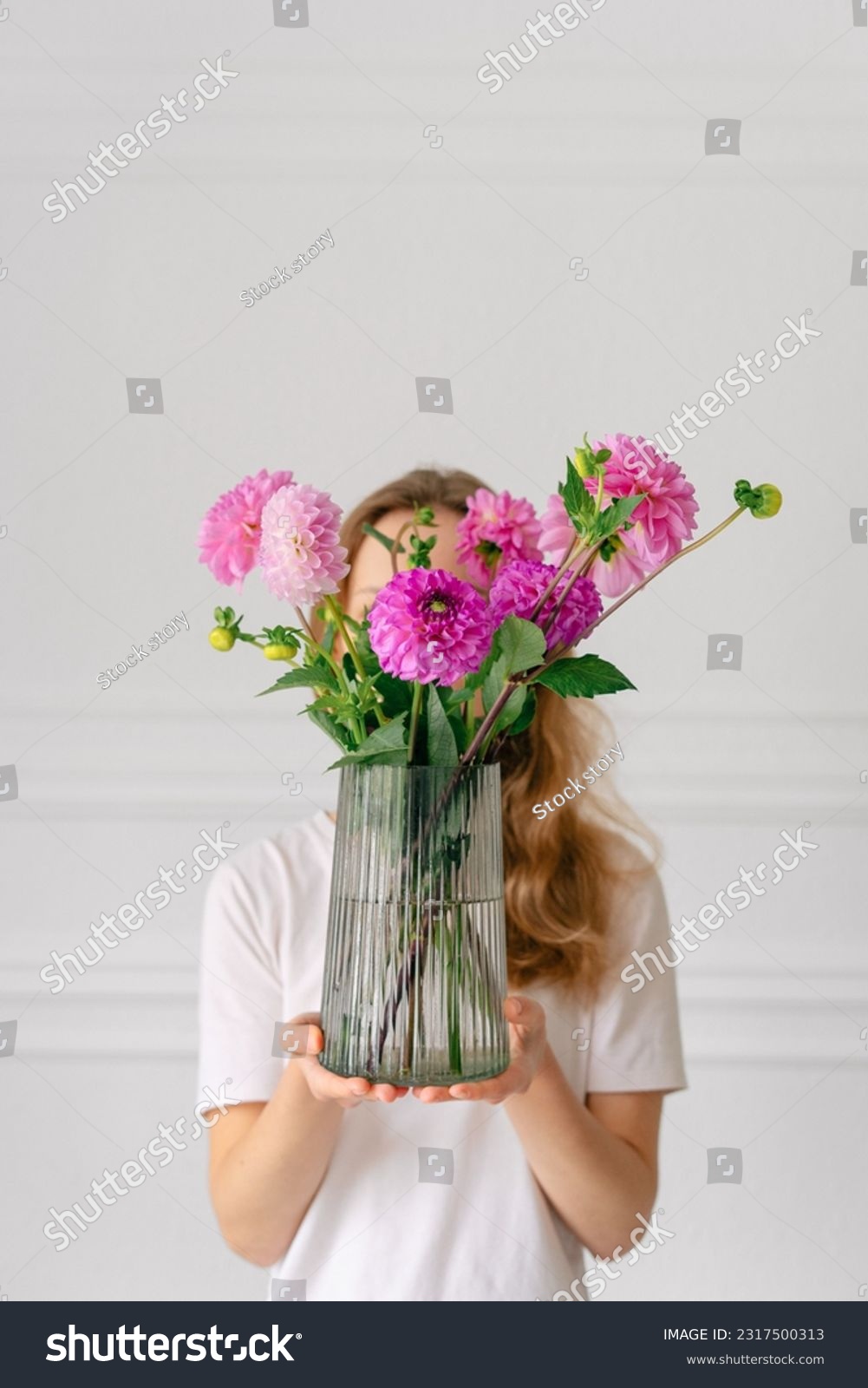 Vertical image of a young woman holding a bouquet of pink dahlias in a vase in front of her on a white background. The concept of flower workshops and articles for the study of floristry #2317500313