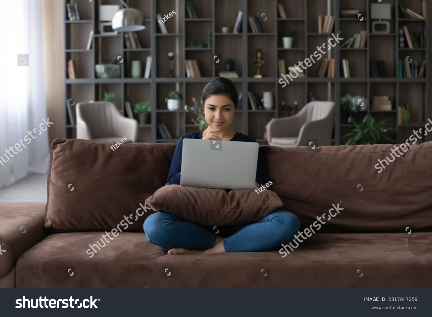 Young indian woman sit cross legged on couch enjoy online date create answer on message from boyfriend at social network. Curious hindu female read interesting information at blog website on pc screen #2317847259