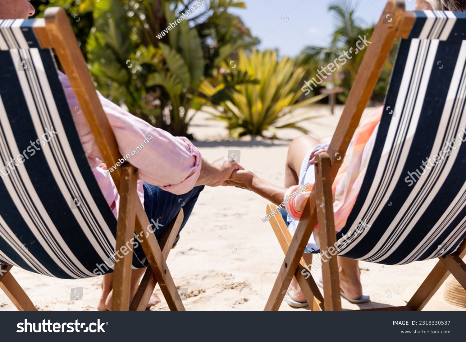 Low section of caucasian romantic senior couple holding hands and sitting on deck chairs at beach. Copy space, unaltered, retirement, vacation, love, together, enjoyment, relaxing nature and summer. #2318330537