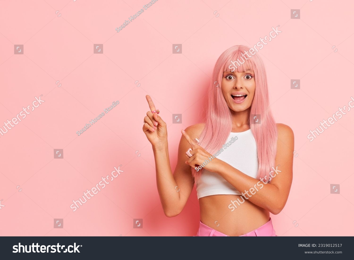 Young pretty woman stands against pink background, pointing index fingers aside with smile, wearing white top, pleasure concept, copy space #2319012517