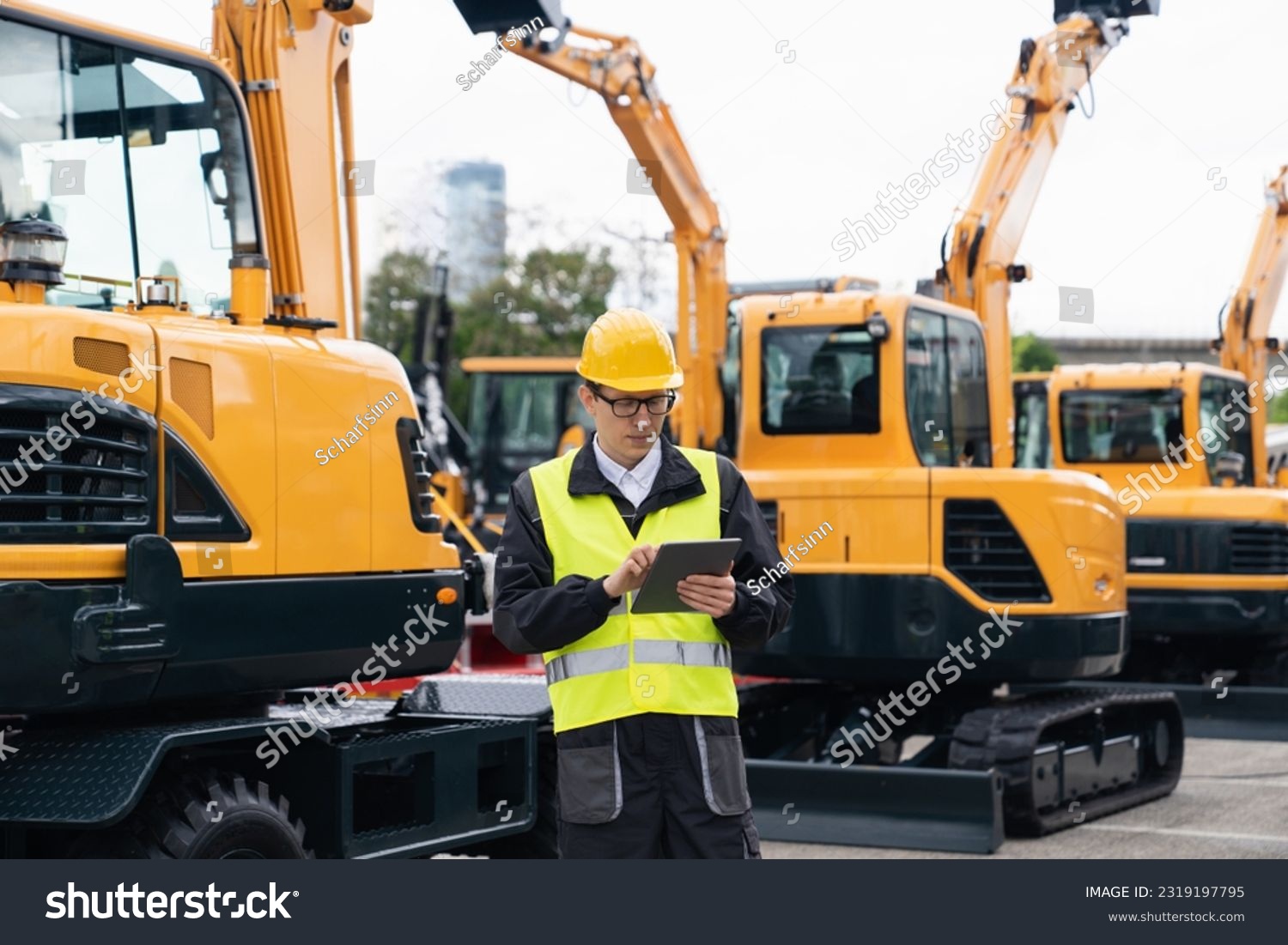 Engineer in a helmet with a digital tablet stands next to construction excavators	 #2319197795